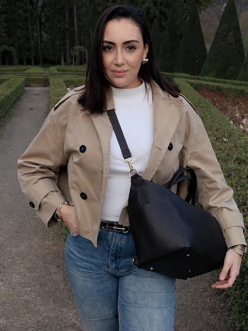 Une cliente en trench porte son sac ALBA noir dans un jardin